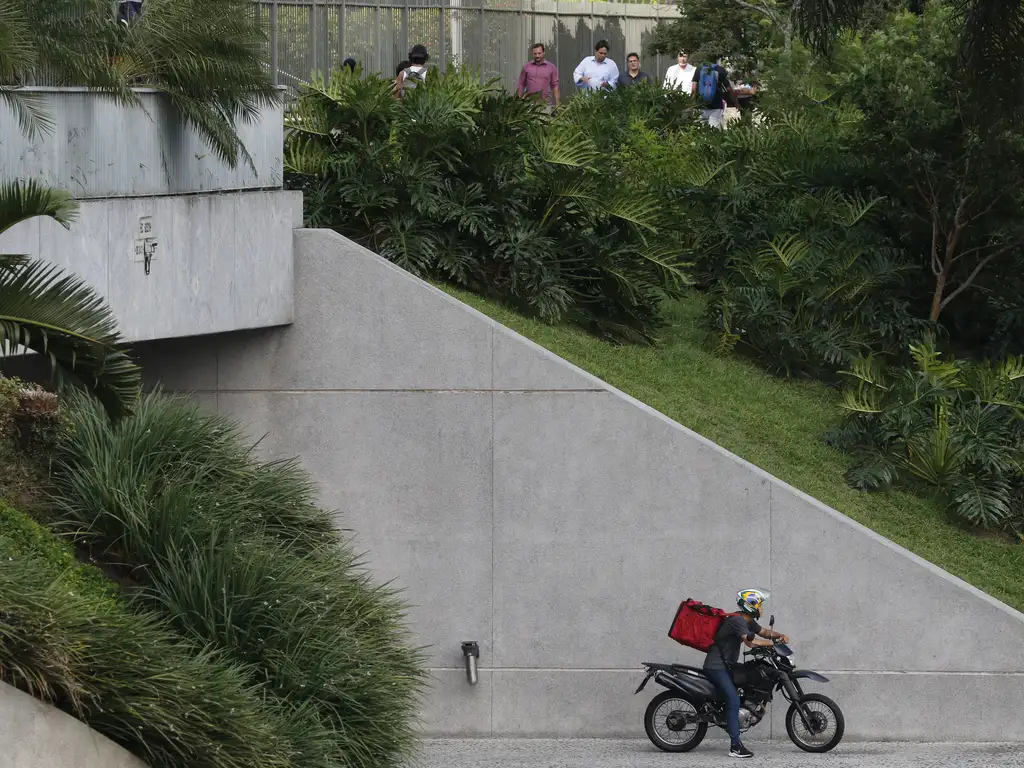 A imagem mostra uma cena ao ar livre com um foco em uma rampa de concreto que leva a um prédio. Na rampa, há uma pessoa pilotando uma motocicleta, usando um capacete e carregando o que parece ser uma mochila vermelha de entrega. O motociclista está se movendo em direção ao canto inferior direito da imagem. Acima e à esquerda da rampa, há uma passarela elevada onde várias pessoas estão de pé atrás de grades, aparentemente observando o ambiente ou esperando. A área é cercada por vegetação exuberante com várias árvores e arbustos, indicando um cenário urbano com esforços de paisagismo.