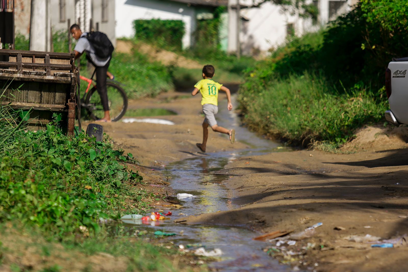 A imagem mostra uma cena ao ar livre com uma criança correndo descalça em um caminho molhado e lamacento. A criança está vestindo uma camisa amarela com o número 10 nas costas. Ao fundo, há um adulto andando de bicicleta e outra pessoa em pé ao lado de algo que parece ser um carrinho ou carroça de madeira. O chão está cheio de lixo e detritos, indicando possível negligência ou falta de serviços de saneamento na área.