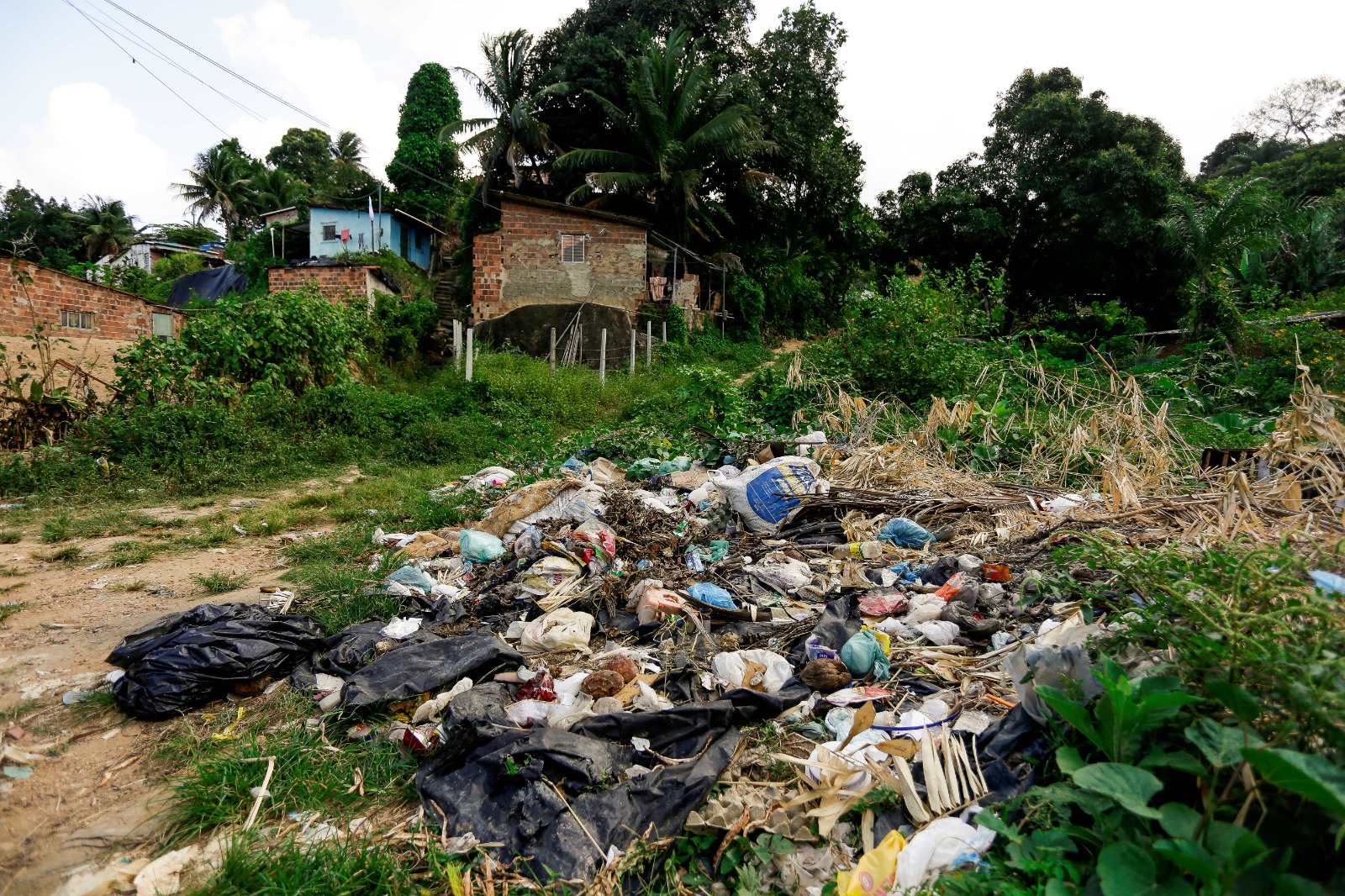 A imagem mostra uma pilha de lixo variado em primeiro plano, com diferentes tipos de resíduos como garrafas plásticas, sacolas e outros itens não identificáveis. Atrás do lixo, há uma área de grama alta e, mais ao fundo, várias casas em uma colina cercada por vegetação densa. O céu está nublado.