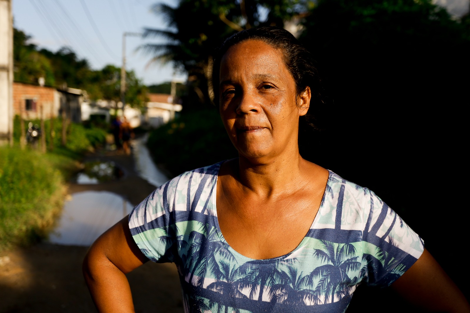 Na foto, há uma mulher negra, de cabelos pretos presos por tráa da cabeça, em primeiro plana. Ela está vestindo uma blusa de mangas curtas com um padrão que inclui tons de azul, branco e verde, lembrando folhas de palmeira ou folhagem. Ao fundo, há uma cena de rua com casas de ambos os lados de uma estrada não pavimentada. Há árvores e vegetação visíveis ao redor das casas, e o céu está claro, com a luz do dia diminuindo, sugerindo que pode ser início da noite ou fim de tarde.