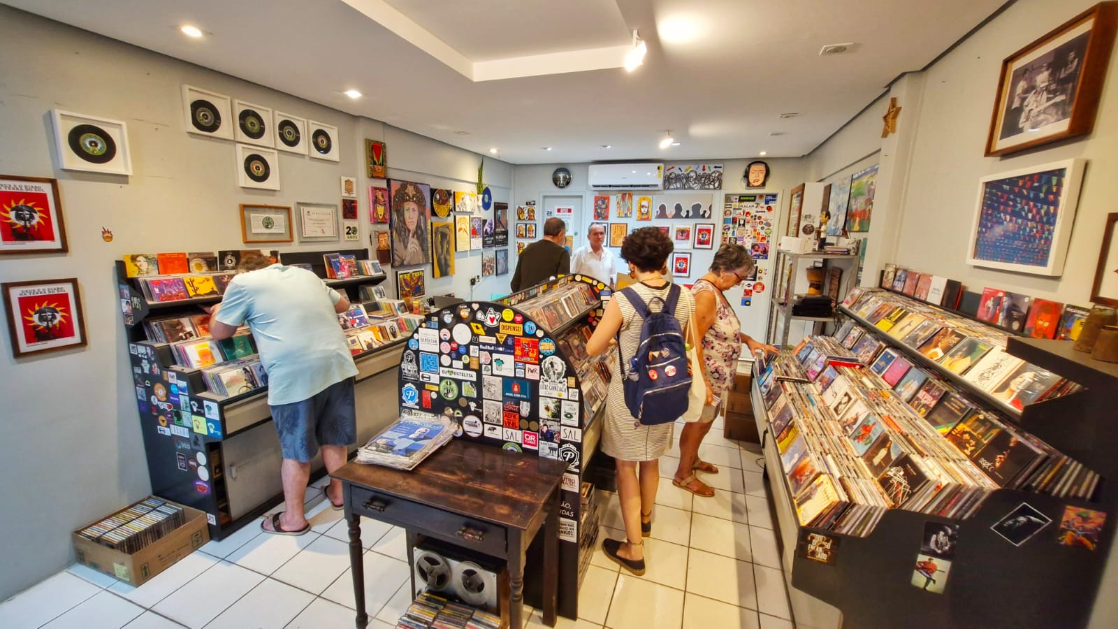 foto mostra o interior de uma loja de discos vibrante e cheia de vida. Há várias prateleiras cheias de discos de vinil, e as paredes estão decoradas com quadros e pôsteres. Vários clientes estão explorando a loja: uma pessoa à esquerda está examinando discos em uma caixa, outra está no centro olhando para uma prateleira, e duas outras estão à direita, uma delas folheando álbuns em uma caixa enquanto a outra está ao lado. O chão é de azulejos brancos e há holofotes iluminando o espaço. A atmosfera sugere um lugar rico em história e cultura musical, provavelmente interessante para entusiastas da música.