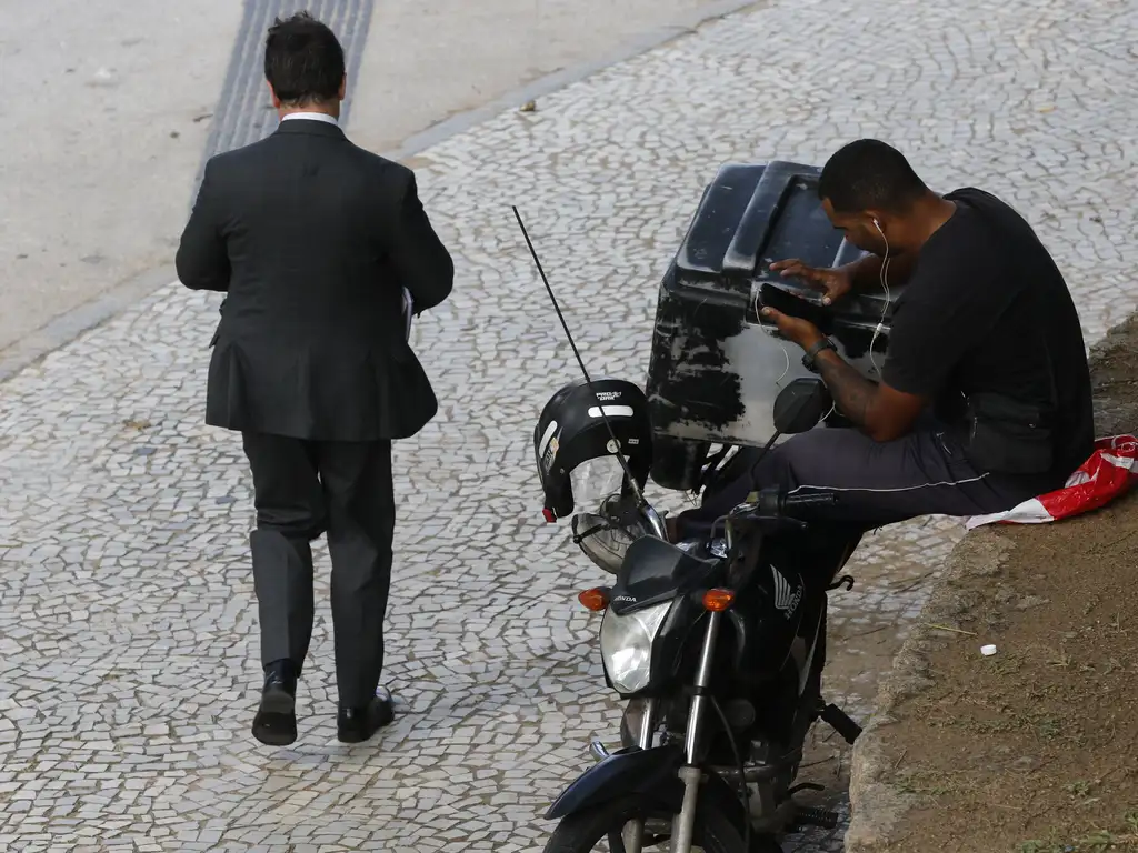 A imagem mostra dois homens em uma rua pavimentada. Um deles está vestida com um terno preto formal e está caminhando de costas para a câmera. A outra pessoa está sentada na calçada ao lado de uma motocicleta estacionada, vestindo roupas casuais e focada em um objeto nas mãos, que pode ser um celular ou um pequeno dispositivo. O contraste entre as roupas e as atividades das duas pessoas pode sugerir estilos de vida ou ocupações diferentes.