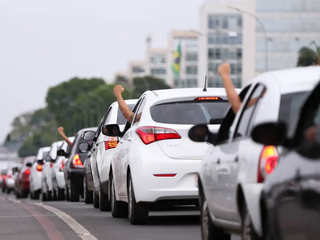 A imagem mostra uma cena de trânsito com uma fila de carros em uma estrada de várias faixas. O foco está na traseira de um carro branco, que se destaca por estar mais próximo e mais nítido em comparação com os outros veículos. O braço de uma pessoa está saindo da janela desse carro, fazendo um gesto de punho fechado, o que pode sugerir frustração ou solidariedade entre os motoristas, embora não seja possível determinar a intenção exata. Ao fundo, há mais carros e alguns prédios urbanos desfocados.