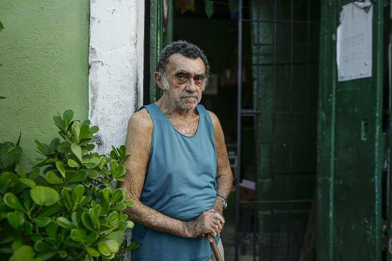 Foto de seu Vital, homem idoso, pardo, de cabelos escuros, costeletas grisalhas e barba mal feita. diante das portas de um prédio antigo apoiado em sua bengala. Ele veste camiseta regata azul.