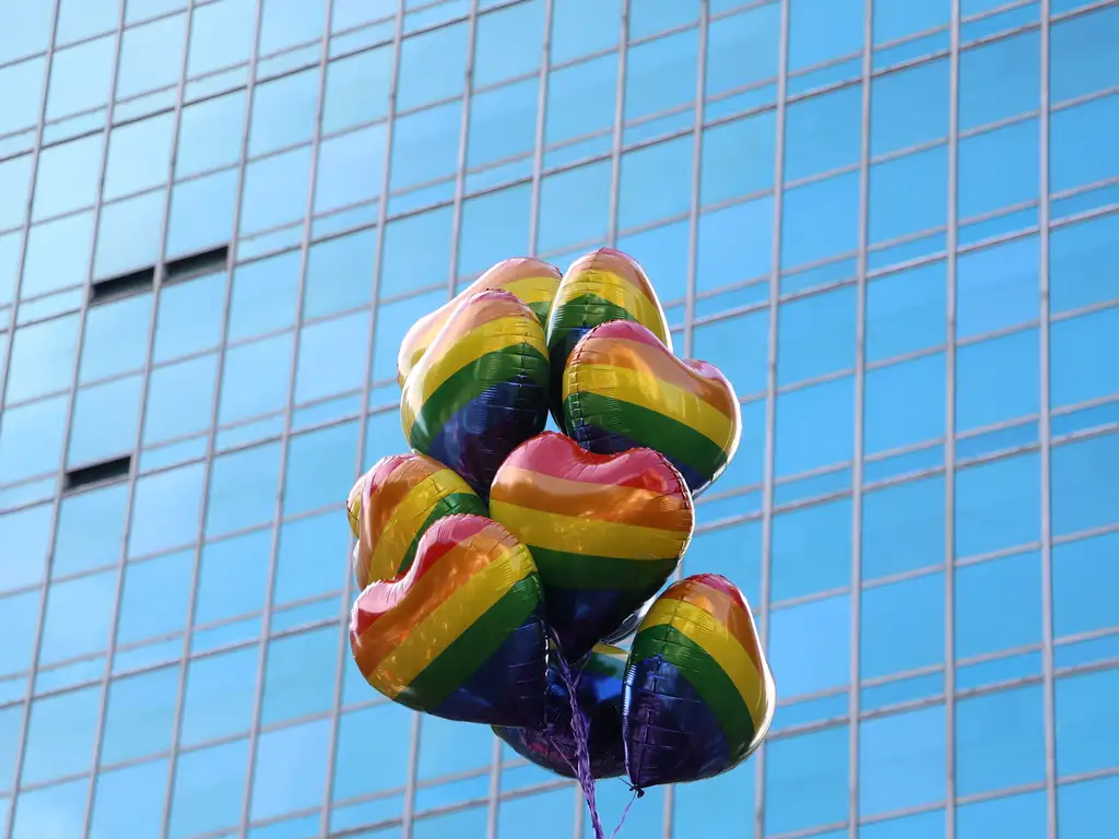 imagem mostra um grupo de balões em forma de coração com um padrão de arco-íris idêntico ao das bandeiras do movimento LGBTQIA+, flutuando em frente a um prédio com janelas de vidro azul.