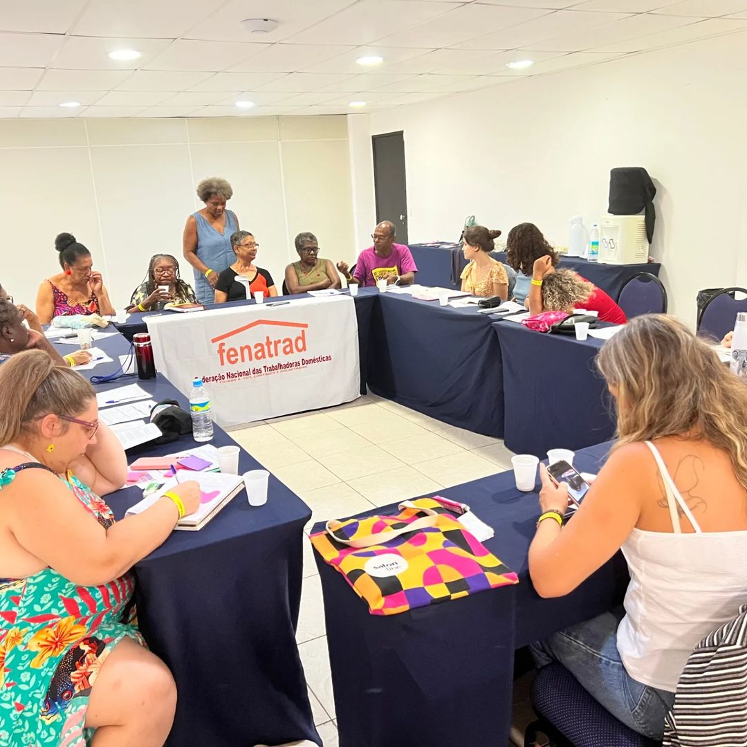 A imagem mostra uma reunião de mulheres sentadas em mesas dispostas em formato de U. No centro, há uma mesa com uma toalha branca que tem o logotipo e o nome fenatrad em vermelho, seguido pela frase Federação Nacional das Trabalhadoras Domésticas. As pessoas estão envolvidas em atividades como escrever, conversar e usar dispositivos móveis. Há copos descartáveis e garrafas de água sobre as mesas. No fundo, há um bebedouro e uma garrafa térmica sobre uma mesa.