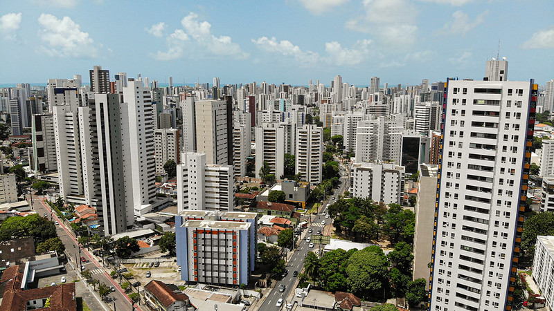 Foto aérea de aglomerado de prédios da zona norte do Recife. na foto, se vê a linha do horizonte tomada por prédios, de vários tamanhos, e o céu azul no topo. O céu está azul, com poucas nuvens.