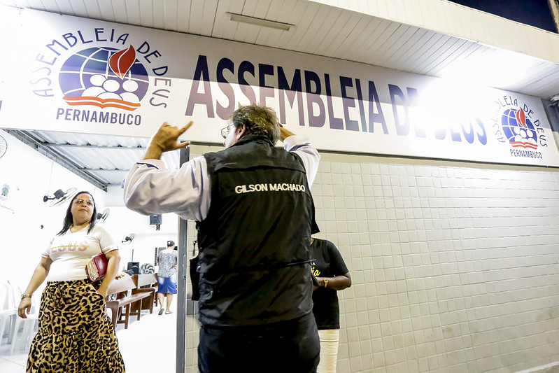 A imagem mostra a fachada branca de um prédio iluminado com uma grande placa que diz “ASSEMBLEIA DE DEUS” em letras brancas e negrito sobre um fundo azul. Abaixo, há outro letreiro indicando “PERNAMBUCO”. Uma mulher está saindo do prédio, olhando de lado para um homem que está na frente do local, com os bracos agitados e usando um colete preto com o nome “GILSON MACHADO” visível nas costas. 