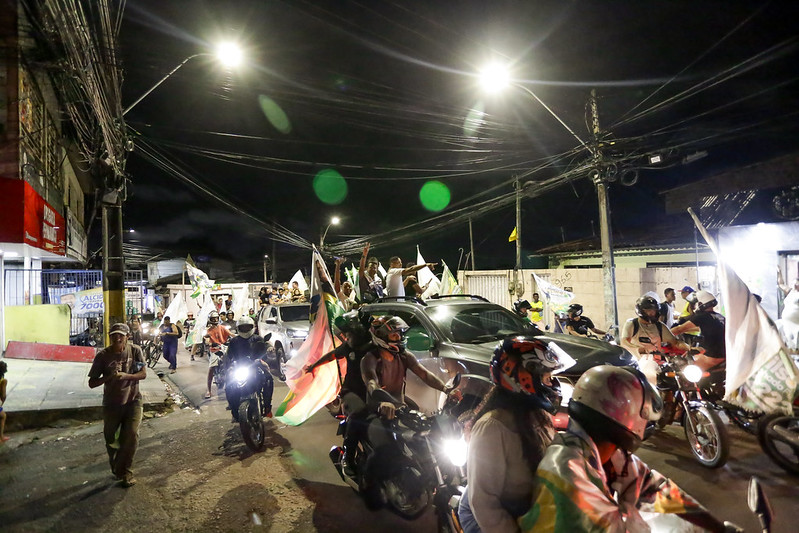 A foto mostra uma cena noturna de rua com um grupo de pessoas, algumas a pé e outras em motocicletas. Há vários cabos elétricos cruzando acima da rua. As pessoas nas motocicletas estão carregando bandeiras de campanha eleitoral. A cena sugere um ambiente animado que pode estar associado a uma celebração ou reunião pública. Há iluminação artificial de postes de luz e faróis que iluminam partes da cena.