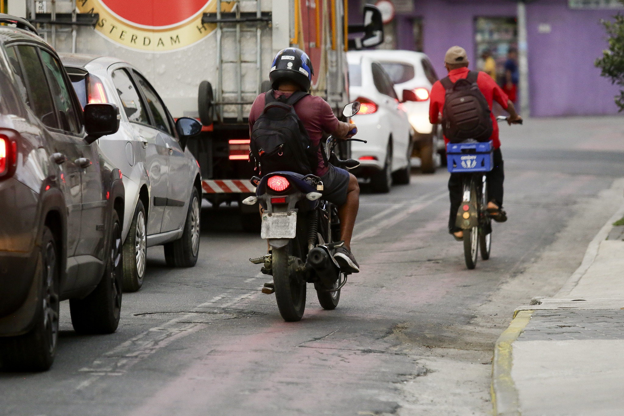 A imagem mostra uma cena urbana com vários tipos de veículos em uma rua movimentada. Além de carros, há uma motocicleta e uma bicicleta transitando por uma ciclofaixa. A motocicleta é conduzida por uma pessoa usando capacete, mochila e camisa roxa, enquanto a bicicleta também é conduzida por um homem com mochila preta, usando camisa vermelha, calça preta e um boné bege. Há um caminhão grande à frente. No fundo, vemos edifícios e uma parede roxa.