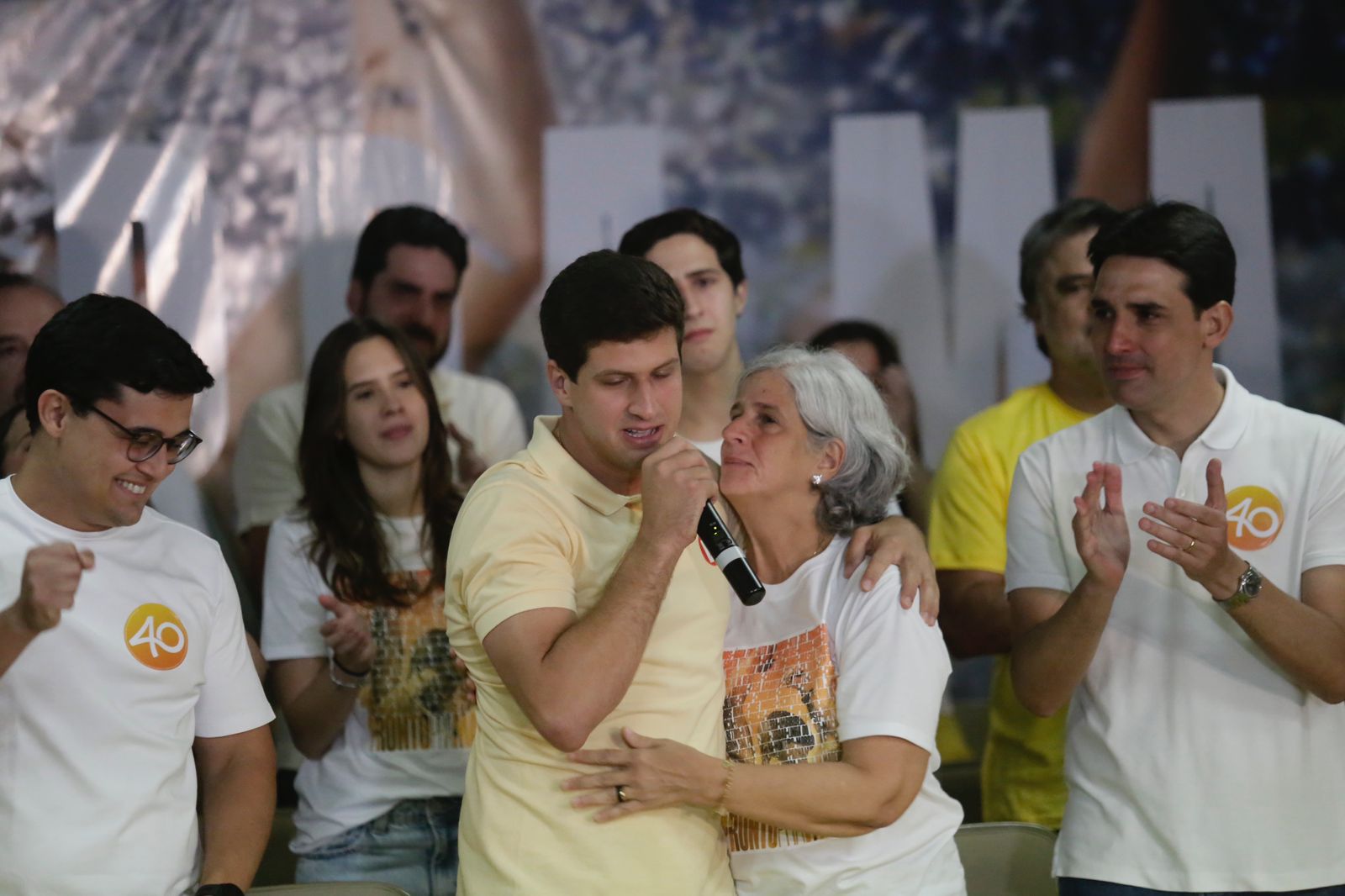 Foto de João Campos, prefeito reeleito do Recife, homem branco e jovem, usando camisa polo amarelo claro, abraçado com sua mãe, Renata Campos, mulher branca, de cabelos brancos. Ambos estão chorando, visivelmente emocionados, sendo observados por um grupo de pessoas brancas, usando roupas claras.