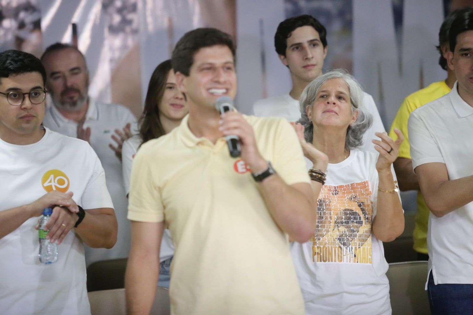 Foto de João Campos, prefeito reeleito do Recife, usando camisa polo amarela clara, entre sua mãe Renata - usando camiseta branca - e o vice-prefeito Vitor Marques, homem branco, jovem, de óculos e camisa branca. Ao fundo, algumas pessoas o observam enquanto ele fala ao microfone.