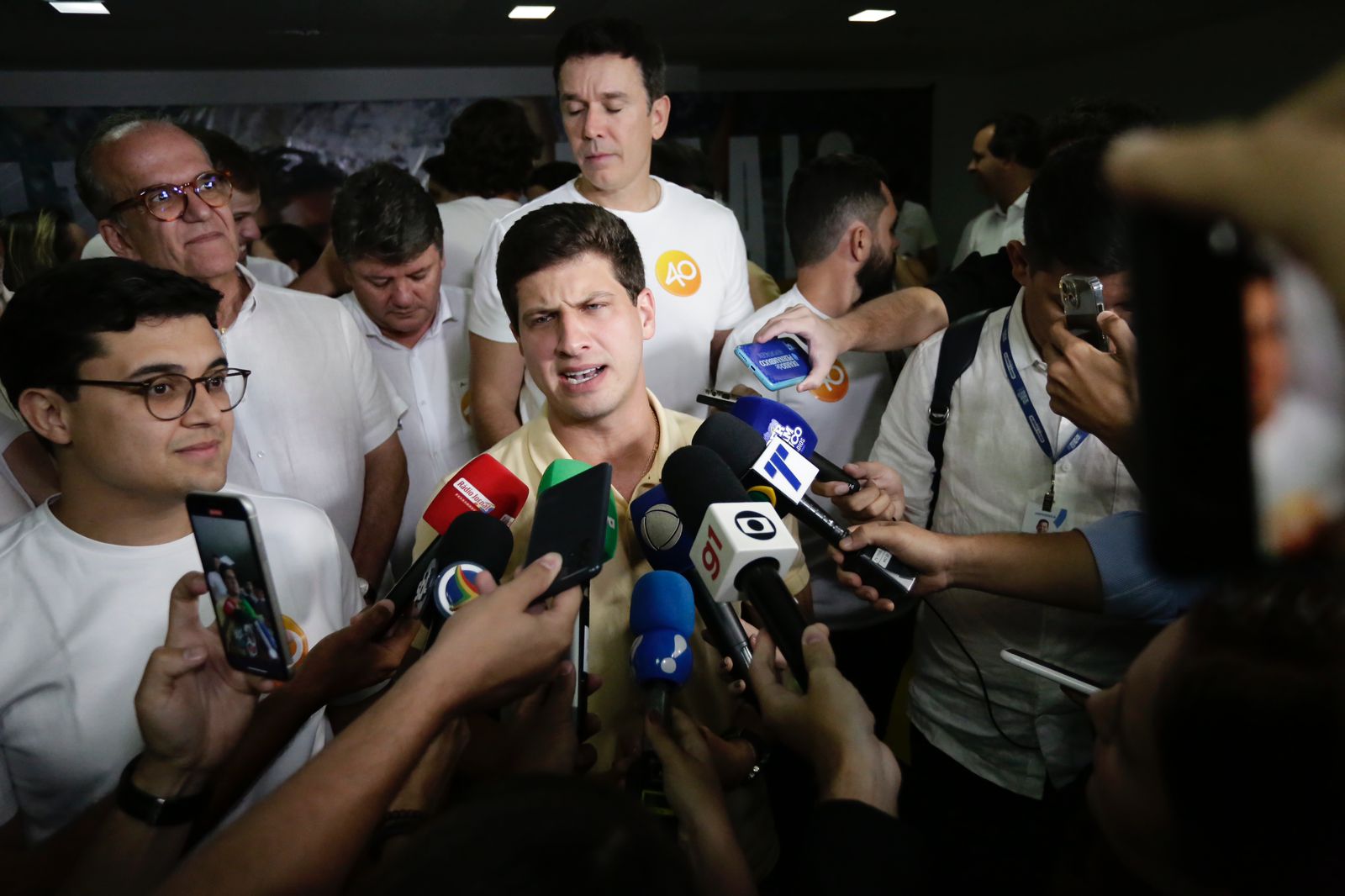 Foto de João Campos dando entrevista cercado por microfones, tendo à esquerda o vice-prefeito Vitor Marques, homem branco, jovem, de óculos de aros redondos. Ao redor do grupo de entrevistados e repórteres há várias pessoas usando roupas claras. A foto foi feita em um ambiente fechado.