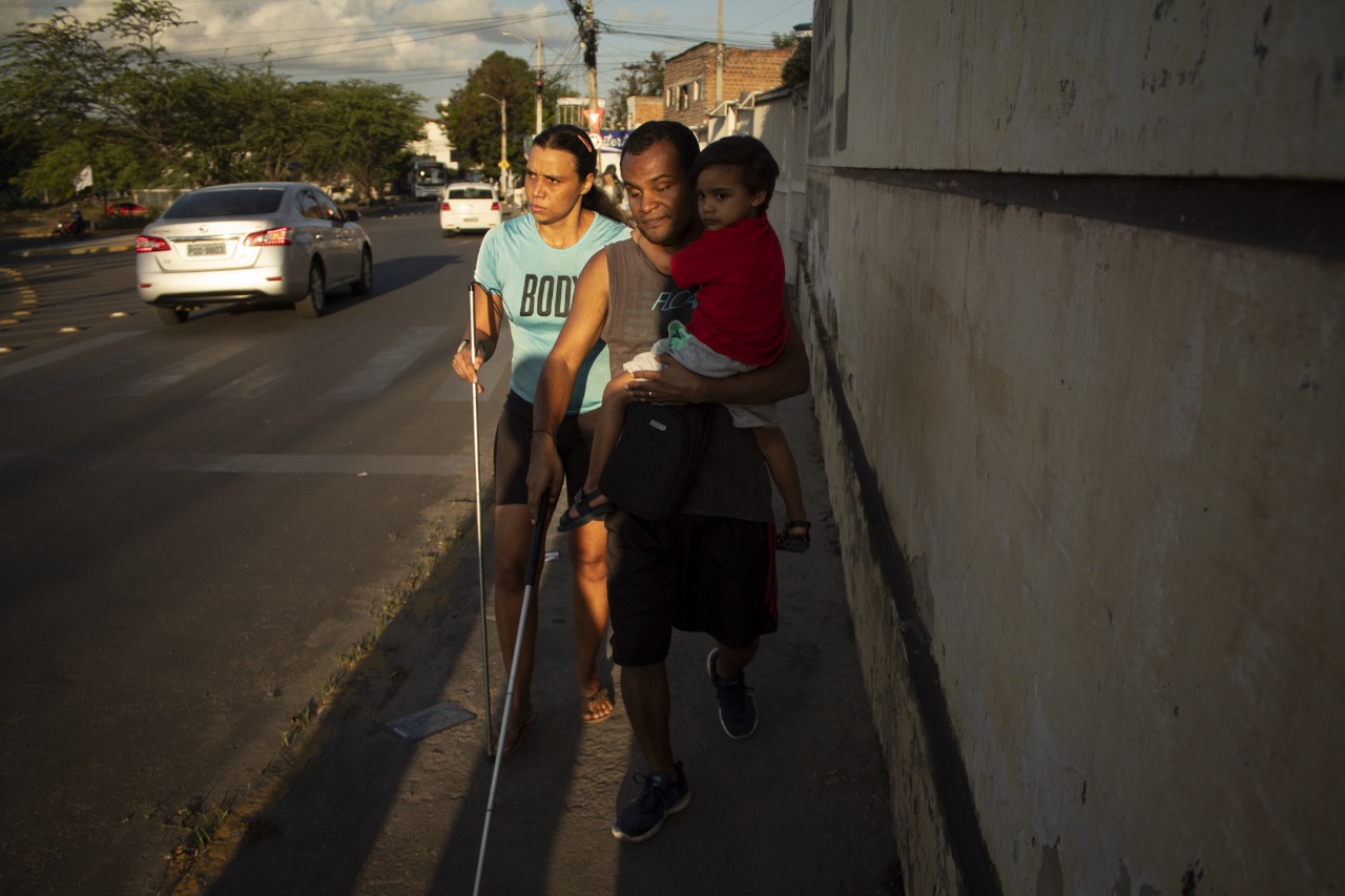 A imagem mostra um casal caminhando em uma calçada ao lado de uma rua com carros em movimento. O homem negro em primeiro plano está usando uma bengala branca, indicando deficiência visual, e levando no colo um menino com camiseta vermelha A mulher ao fundo, à esquerda da imagem, veste uma camiseta verde claro e também usa uma bengala branca. Ao fundo, há prédios e parece ser de dia, pois a luz do sol projeta sombras no chão.