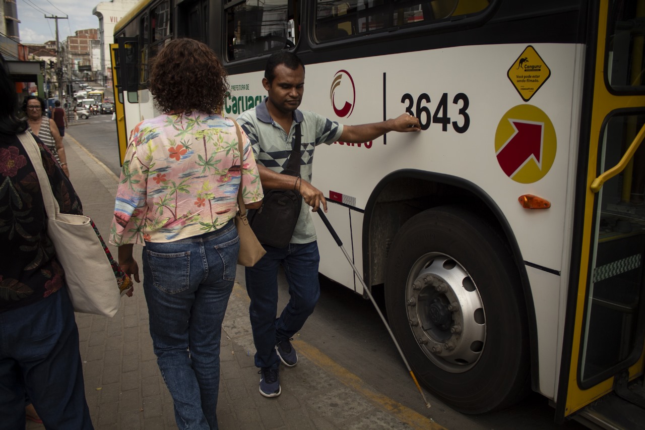 A imagem mostra uma cena urbana com um ônibus de transporte público e várias pessoas. O ônibus é branco com gráficos vermelhos e amarelos, incluindo um logotipo circular com centro vermelho e borda branca, e o número 3643 destacado. Um homem cego usando uma bengala e vestindo uma camisa cinza está embarcando no ônibus. O cenário parece ser um ponto de ônibus com outros pedestres por perto.