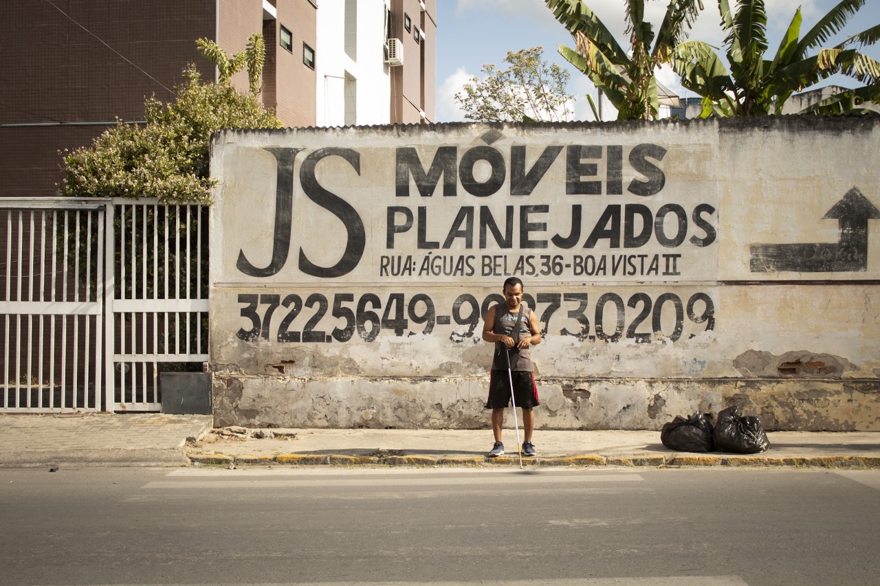 A imagem mostra uma cena urbana ao ar livre com um homem negro cego em pé, tentando atravessar uma rua em frente a uma parede que tem um anúncio pintado. A parede faz parte de uma estrutura maior que parece ser um prédio comercial ou residencial. Há também uma cerca de metal à esquerda da pessoa e alguns sacos de lixo no chão perto dela. O céu está claro, sugerindo que pode ser um dia ensolarado.