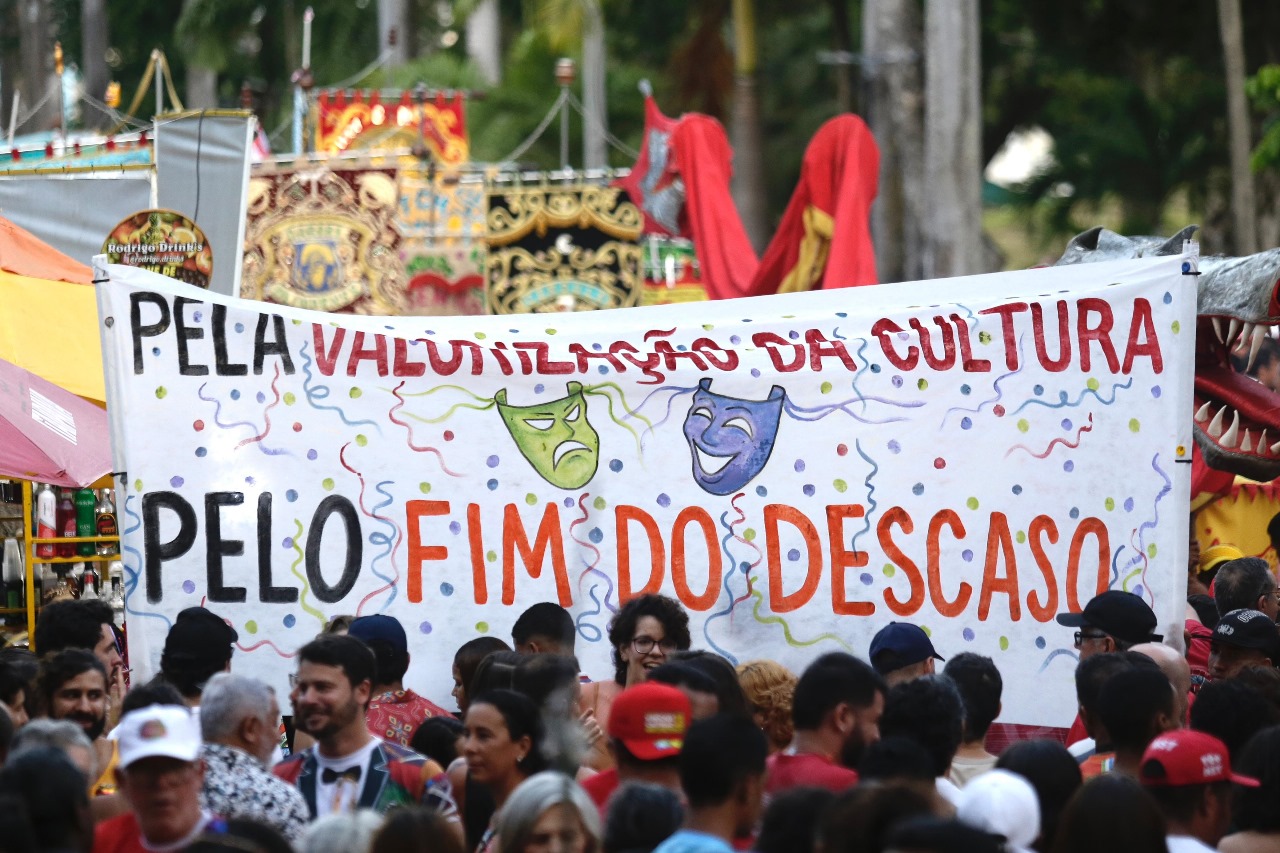 Foto colorida que mostra centenas de foliões reunidos no Sítio Histórico de Olinda. A foto mostra uma grande faixa onde está escrito Pela valorização da cultura, pelo fim do descaso. Atrás, aparecem diversos estandartes de agremiações do frevo.