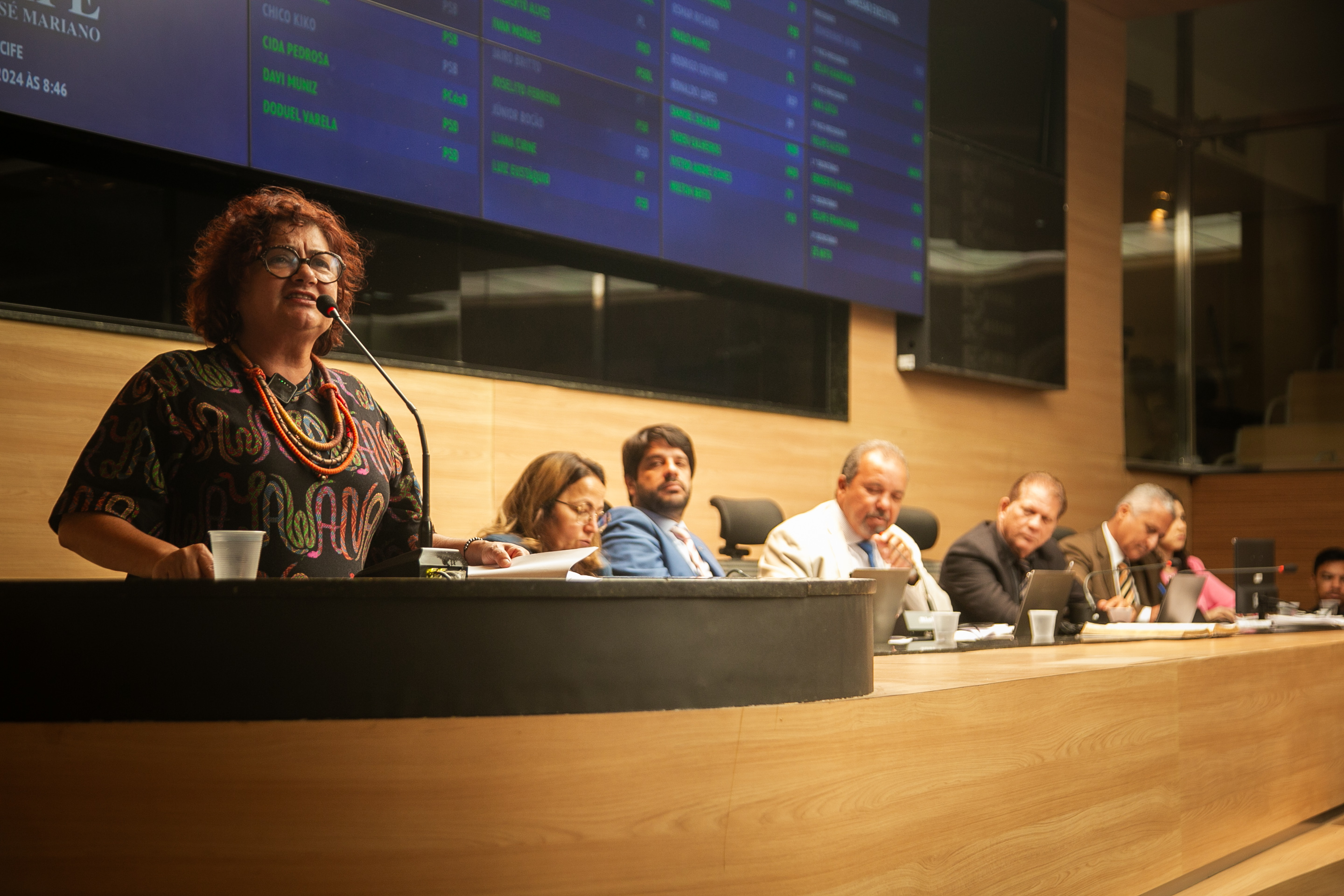 A imagem mostra uma sessão em um plenário, onde várias pessoas estão sentadas em uma mesa longa de madeira clara. Uma mulher, Cida Pedrosa, está em pé, falando ao microfone, enquanto outras estão sentadas, algumas olhando para papéis ou dispositivos eletrônicos. Cida Pedrosa tem cabelos ruivos, óculos de armação redonda, vestido escuro estampado com grafismos laranjas e amarelos, além de um colar laranja e amarelo que dá três voltas em seu pescoço. No fundo, há um grande painel eletrônico com nomes e números, possivelmente indicando votos ou presença de membros. A sala tem um design moderno, com paredes de madeira clara e iluminação suave.