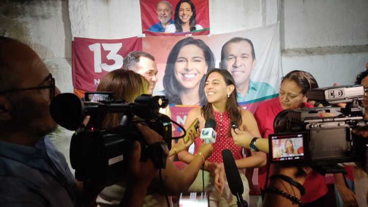 A imagem mostra uma coletiva de imprensa. No centro da imagem, há uma mulher branca, de cabelos escuros compridos. É a candidata a prefeita de Natal, Natália Bonavides. Ela está usando uma blusa vermelha com bolinhas brancas, cercada por várias pessoas segurando microfones e câmeras. Ao fundo, há um banner com o número “13” e fotos com rostos de pessoas, incluindo a própria Natália. A foto foi feita em um ambiente fechado.