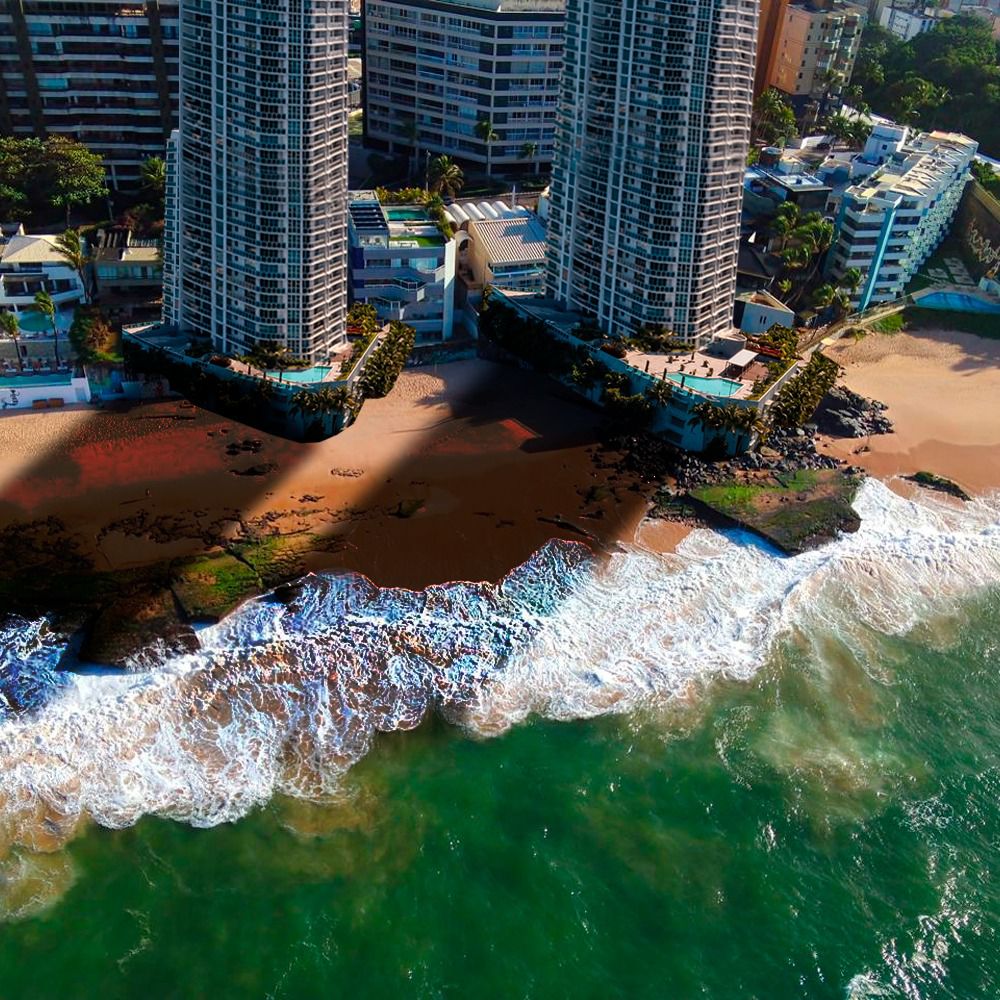 A foto mostra uma vista aérea de uma cena costeira. Há dois prédios altos com várias varandas, situados próximos à praia. Entre esses prédios, há uma área de pátio que leva até a orla. A praia tem areia de cor marrom-avermelhada e é cercada por rochas em ambos os lados, com ondas quebrando contra elas. A água do oceano muda de um tom mais claro perto da costa para um azul mais profundo à medida que se estende para fora, indicando diferentes profundidades.