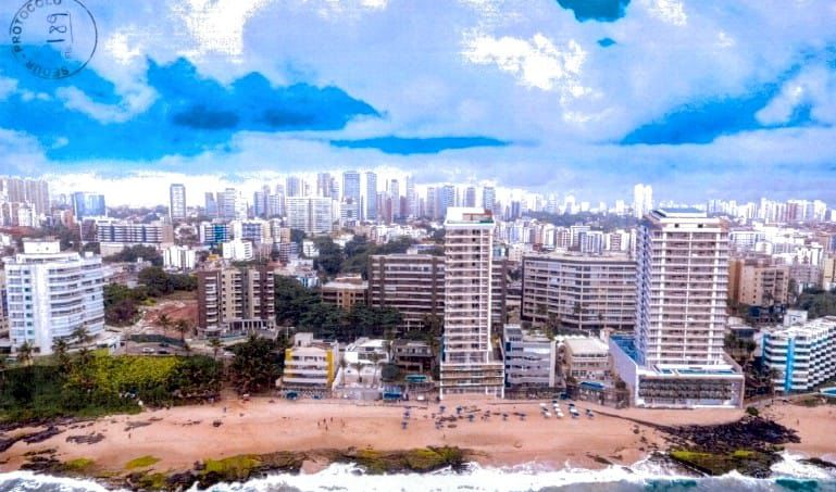 A imagem mostra uma cidade costeira. Há uma praia com ondas quebrando na areia e, além da praia, há uma densa coleção de prédios altos que parecem ser apartamentos residenciais ou hotéis. A arquitetura é variada, mas principalmente de estilos modernos. O céu está parcialmente nublado com algumas áreas azuis visíveis, sugerindo que pode ser um dia ensolarado.