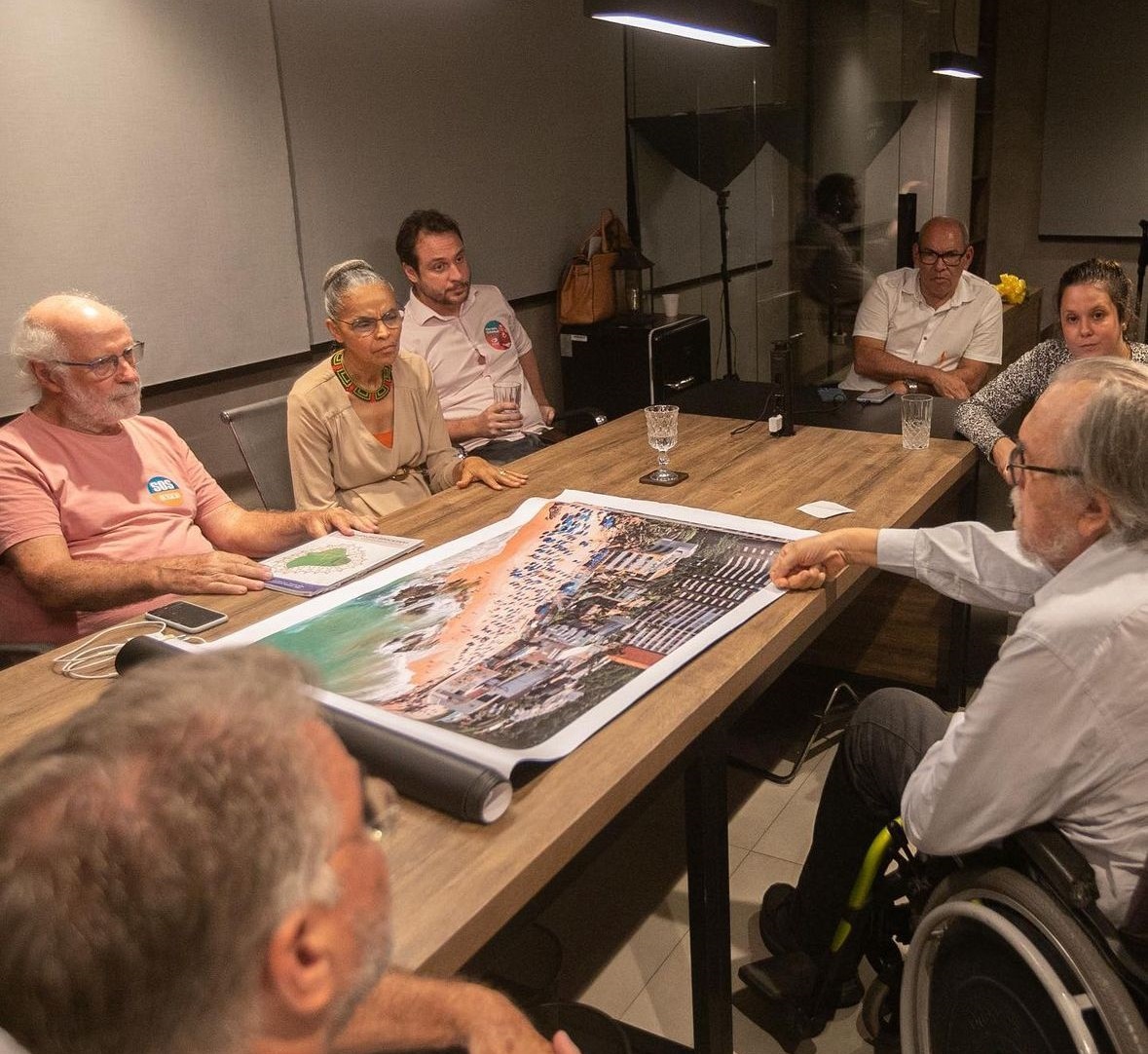 A foto mostra um grupo de pessoas sentadas ao redor de uma mesa, aparentemente em uma reunião ou discussão. No centro da mesa, há uma grande fotografia aérea ou mapa detalhado, que está sendo analisado pelo grupo. Há pelo menos sete pessoas visíveis, tendo ao centro a ministra Marina Silva. Uma das pessoas está sentada em uma cadeira de rodas, indicando um ambiente inclusivo. A sala tem iluminação suave e há um reflexo visível no vidro atrás deles, sugerindo que estão em um ambiente interno