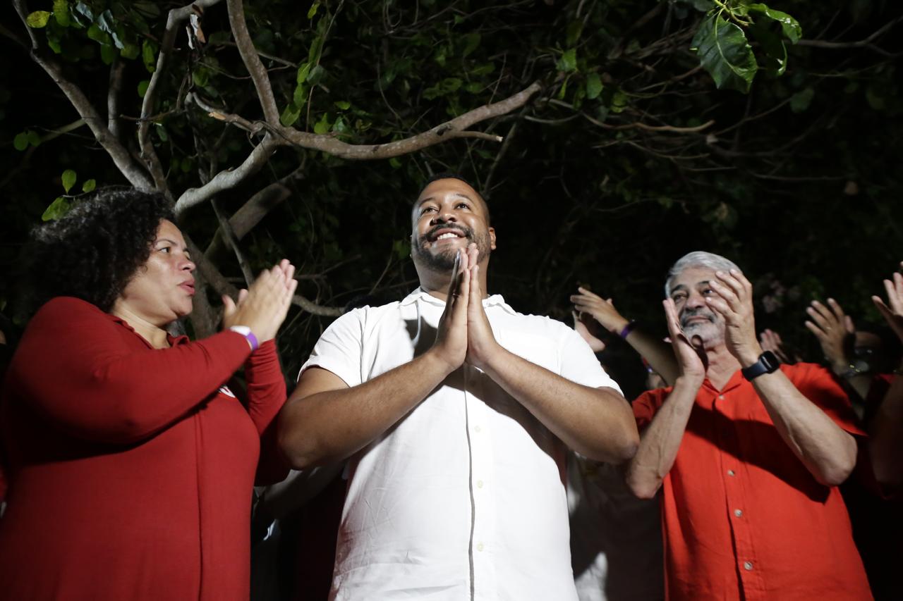 Foto de Vinicius Castello entre Humberto Costa (à direita da imagem) e Dani Portela (à esquerda). em uma foto noturna, sob uma árvore. Ele é um homem homem negro, jovem, de cabelos curtos, barba e bigode ralos, iusando camisa branca, com as mãos postas; Humberto é um homem idoso, branco, de cabelos brancos curtos e bigode e braba grisalhos, usando camisa vermelha, e está aplaudindo; Dani Portela é uma mulher negra, madura, de cabelos cacheados, usando vestido vermelho e que também parece estar batendo palmas.