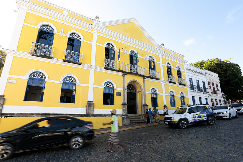 A imagem mostra a fachada da prefeitura de Olinda, um edifício histórico de dois andares, pintado de amarelo com detalhes brancos. Várias janelas têm molduras decorativas e há varandas de ferro forjado. Na fachada, há três bandeiras hasteadas. A rua de paralelepípedos à frente do edifício tem alguns carros estacionados e uma pessoa caminhando. O céu está claro, indicando um dia ensolarado. O edifício ao lado é branco com detalhes em azul e vermelho.