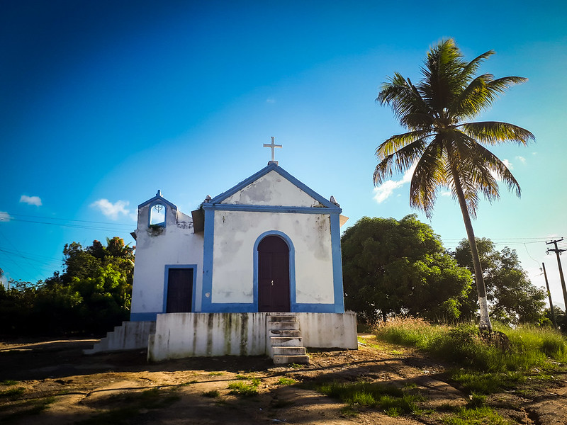 Foto de uma pequena igreja católica pintada de branco com detalhes em azul e encimada por uma cruz branca. A pintura mostra sinais de degradação, com manchas de umidade nas paredes. A igreja foi construída sobre uma plataforma ou patamar em um terreno descampado tendo árvores de grande porte ao fundo e um coqueiro à direita. O dia está claro com o céu azul sem nuvens.