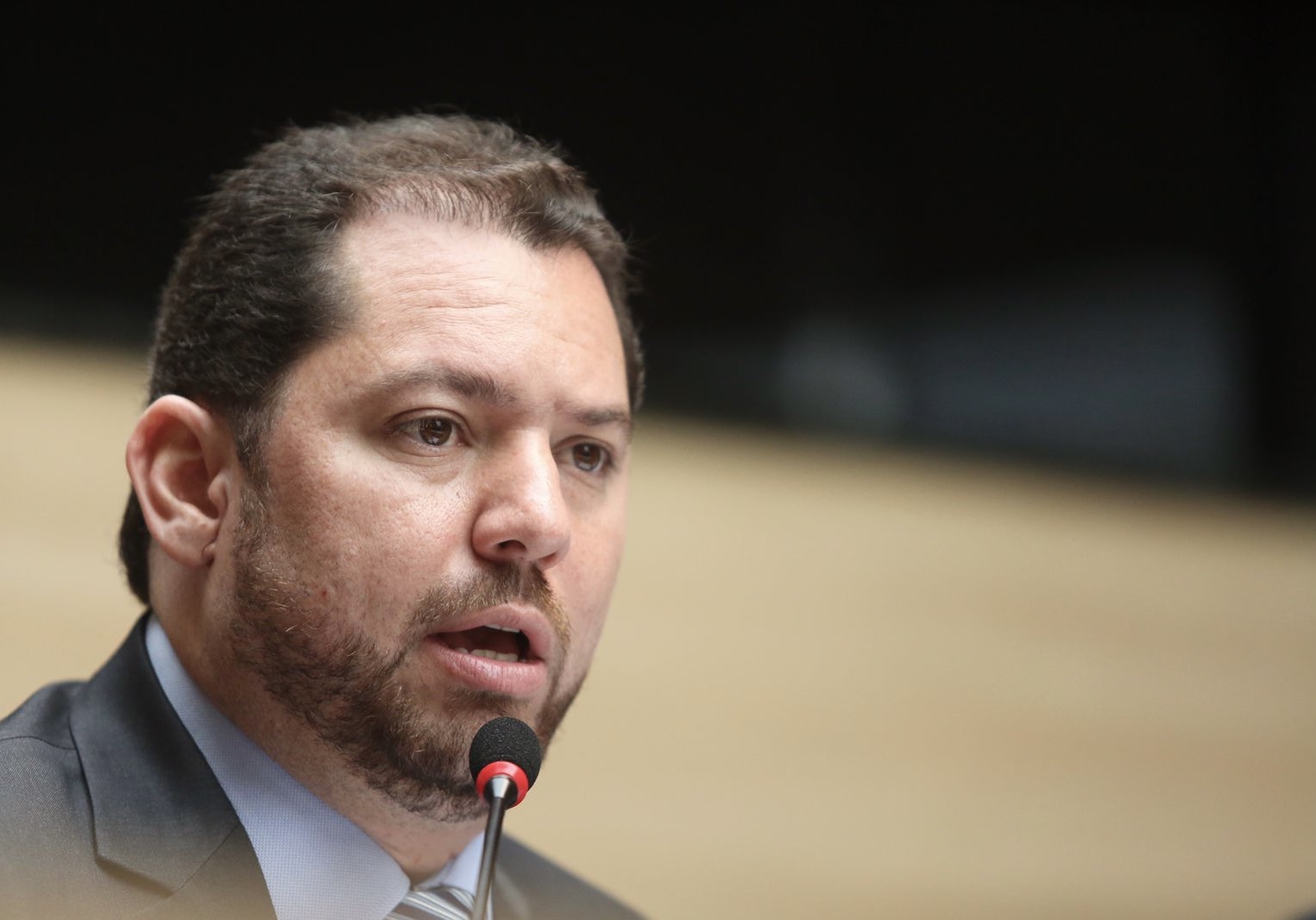 Foto em close de Romerinho Jatobá. Ele é um homem branco, de barba rala castanha e cabelos escuros, com calva pronunciada. Ele está falando ao microfone tendp ao fundo um cenário desfocado.