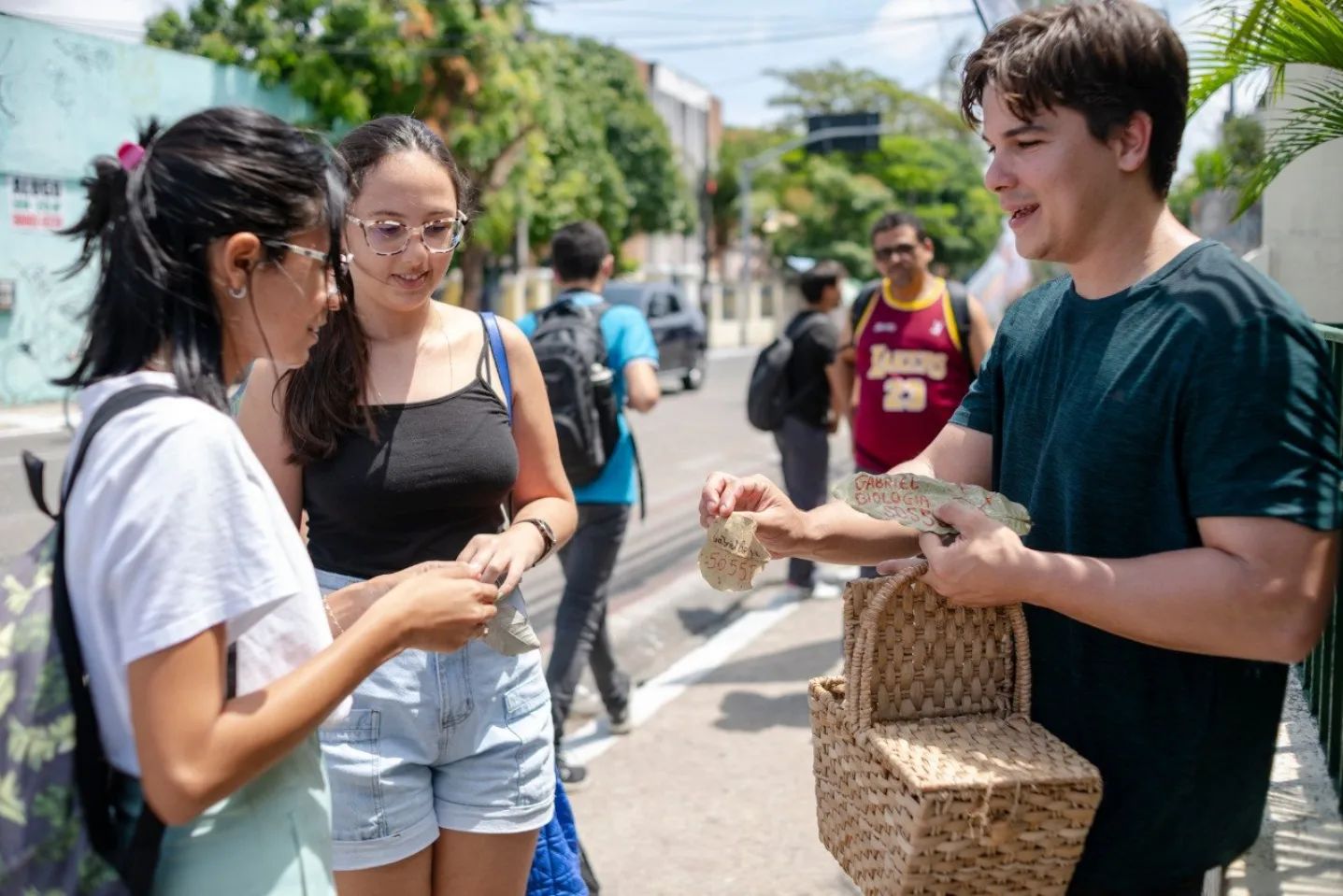 Foto de Gabriel Aguiar, homem branco, de cabelos escuros e curtos, vetindo camisa verde, com uma cesta de palha na mão de onde retira folhas secas com seu nome para distribuir a eleitores. Ele está na calçada de uma rua entregando as folhas para duas mulheres jovens, sendo observado por um homem desfocado, com uma camiseta bordô e amrela onde está escrito Lakers 23.