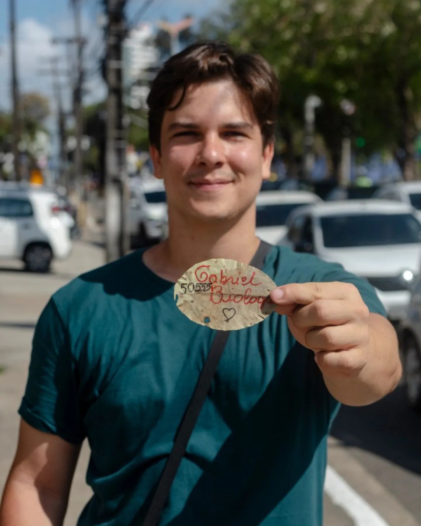 Homem branco jovem, de cabelos pretos curtos, usando uma camisa verde, posando para a câmera exibindo uma folha de árvore seca com o nome Gabriel Biologia e o número 50555. Ele foi fotografado ao ar livre, no meio de uma avenida, com um poste de iluminação pública, árvores e vaarios carros estacionados compondo o cenário ao fundo.