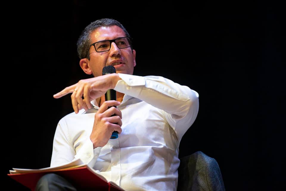 Foto do urbanista e referência na morfologia urbana Vítor Oliveira. É um homem branco de cabelo preto curto e óculos, de meia idade. Na foto, ele está sentado, de camisa branca, e gesticula com um braço e segura um microfone com a outra mão.