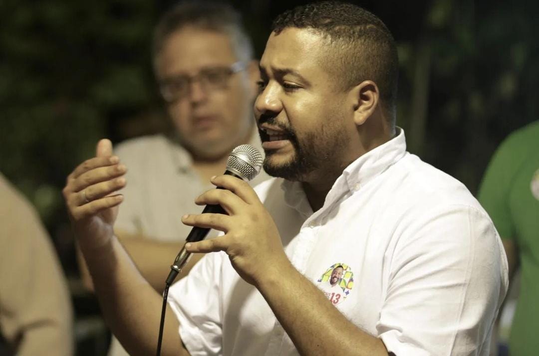 Foto de Vinicius Castello discursando ao microfone em foto noturna. Ele é um homem negro, jovem, de cabelos curtos, vestindo camisa branca, que gesticula ao falar. Enquanto fala, é observado pelo seu candidato a vice, Celso Muniz, um homem branco, de meia idade, cabelos escuros com mechas brancas, cuja imagem está desfocada ao fundo.