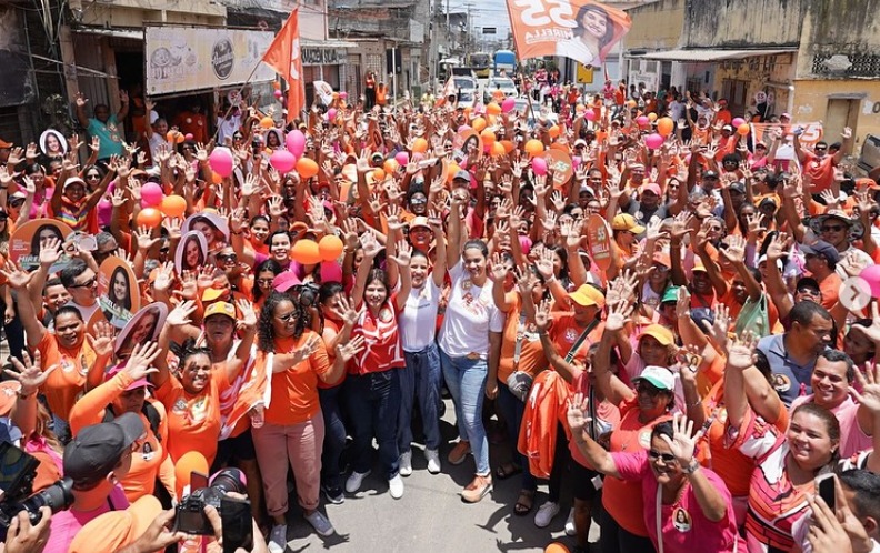 Foto de centenas de pessoas no meio da rua, a maioria usando camisetas laranja, posicionadas para a câmera. Na multidão se sobressaem duas bandeiras laranjas, estampadas com uma foto de mulher branca e o número 55.