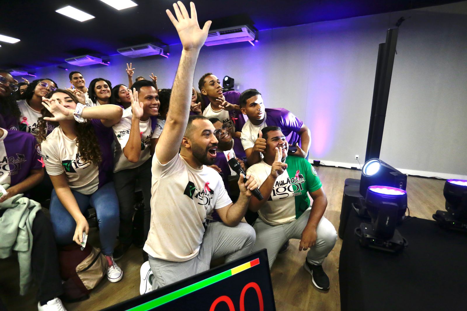 Foto de um grupo de pessoas jovens, diantes de canhões de luzes coloridas, posando para uma câmera que não aparece no vídeo. São rapazes e moças, a maioria pessoas negras, que estão celebrando algo com as mãos levantadas. À frente do grupo, ajoelhado com um microfone na mão, está Gil do Vigor, homem negro, de pele clara, cabelos pertos curtos e barba preta. Ele esta usando uma camisa rosa claro, estampada com palavras de letras pretas ilegíveis causa da posição do corpo.