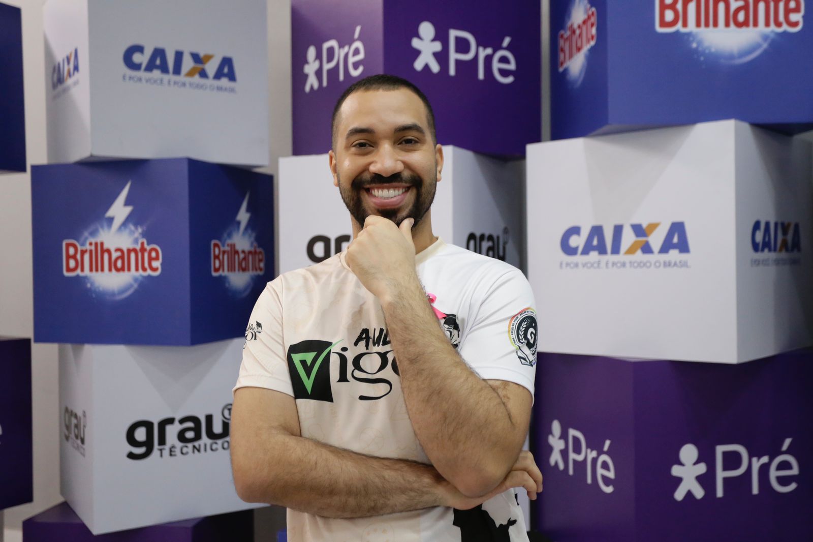 Foto de Gil do Vigor, homem negro, de pele clara, cabelos pertos curtos e barba preta. Ele foi fotografado da cintura para cima com uma camisa roda claro, cujas palavras estampadas em letras pretas estão parcialmente cobertas pelo braço esquerdo dele, cuja mão está tocando o queixo. Ele está rindo para a câmera e, ao fundo, se vê um painel de cubos azuis e brancos onde se lê nomes e marcas de patrocinadores: Caixa, Brilhante, Grau Técnico e Pré, este último acompanhado de um boneco estilizado de figura humana.