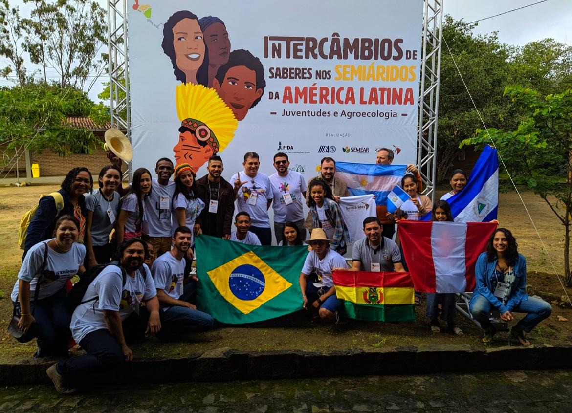 Foto de um grupo de pessoas posando para a câmera segurando bandeiras do Brasil, da Bolívia, Peru, Argentina e Nicarágua. Ao fundo, há um painel branco com a frase Intercâmbio de saberes nos semiáridos da América Latina - Juventudes e Agroecologia ao lado de desenhos de rostos de mulheres e jovens.