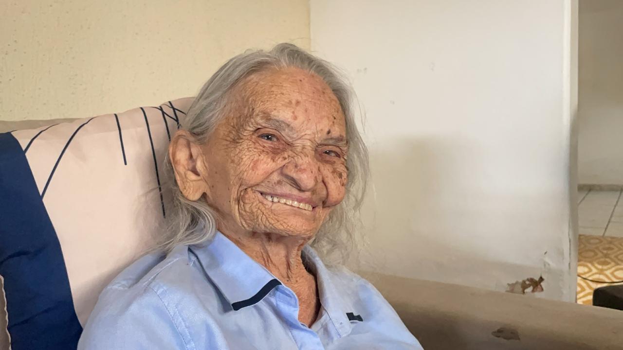 Foto de Elizabeth Teixeira, mulher idosa, de cabelos ralos, rsoto enrugado e pele parda. Ela foi fotograda do tórax para cima vestindo uma bluza azul clara e olhando diretamente para a câmera, sorrindo discretamente.