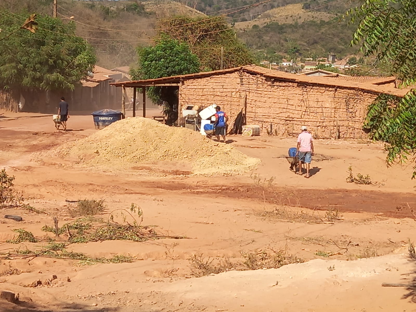 Foto de três homens empurrando carrinhos de mãe com garrafões em um ambiente árido, ao lado de uma casa de paredes de taipa. Na área central da imagem, o solo não tem vegetação e tem cor avermelhada. Ao fundo há árvores esparsas e colinas.