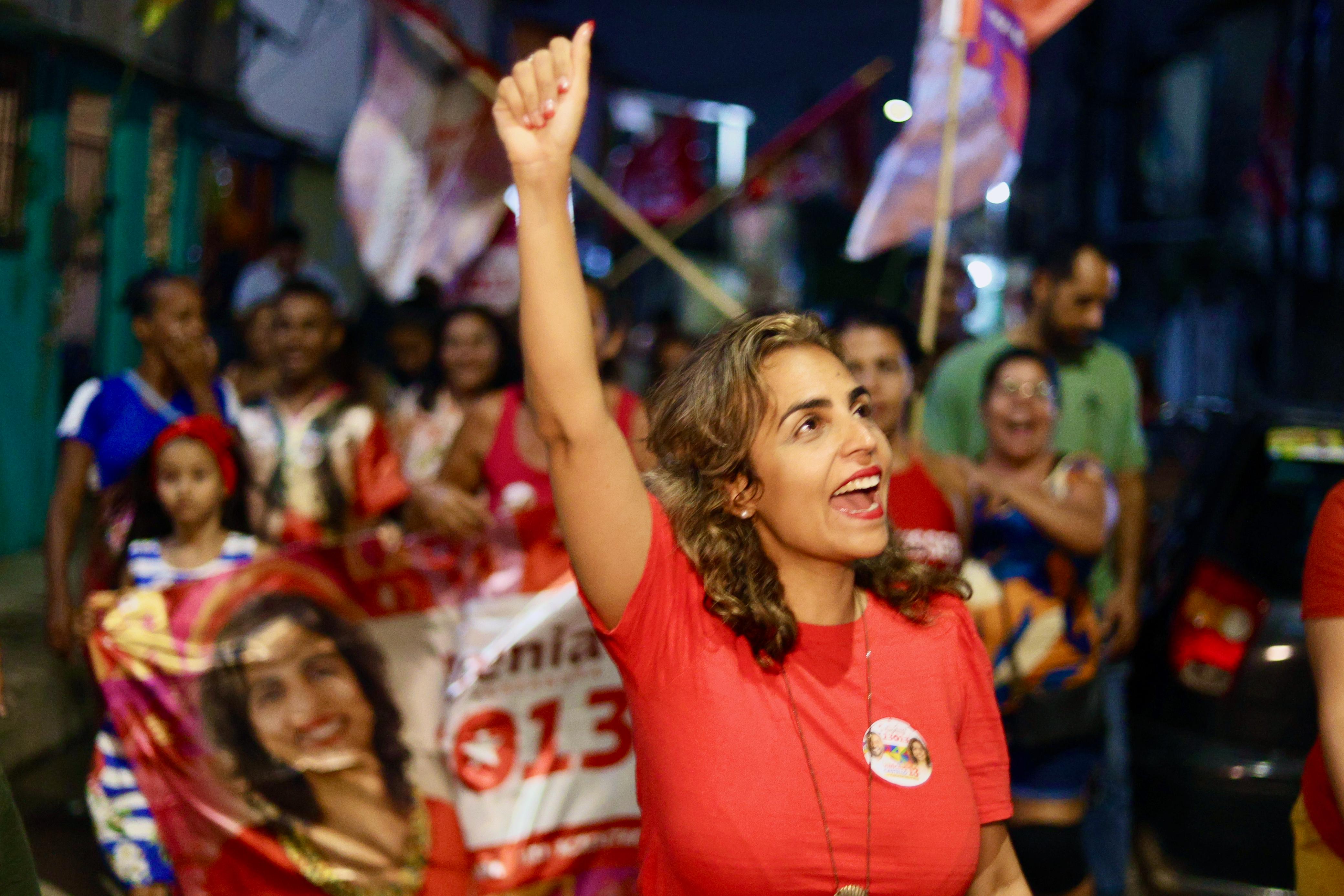 Foto de Eugênia Lima em meio a uma multidão de pessoas vestidas predominantemente de vermelho, com o braço direito erguido e o punho fechado. A foto foi feita ao ar livre e à noite. Eugênia é uma mulher branca, jovem, de cabelos castanhos claros, usando blusa vermelha com um adesivo na altura do peito. Ao fundo há várias bandeiras brancas com letras vermelhas erguidas.