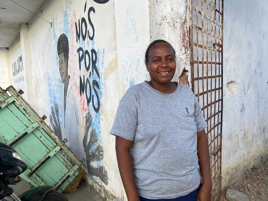 A imagem mostra uma mulher negra, de cabelos presos, em pé em frente a uma parede com arte de grafite. Ela está sorrindo para a câmera, usando uma camiseta cinza. O grafite retrata uma figura com o punho levantado e o texto “NÓS POR NÓS”, sugerindo temas de solidariedade ou autossuficiência. À direita da pessoa, há um portão de metal enferrujado, e à esquerda, parte de uma carroça verde.