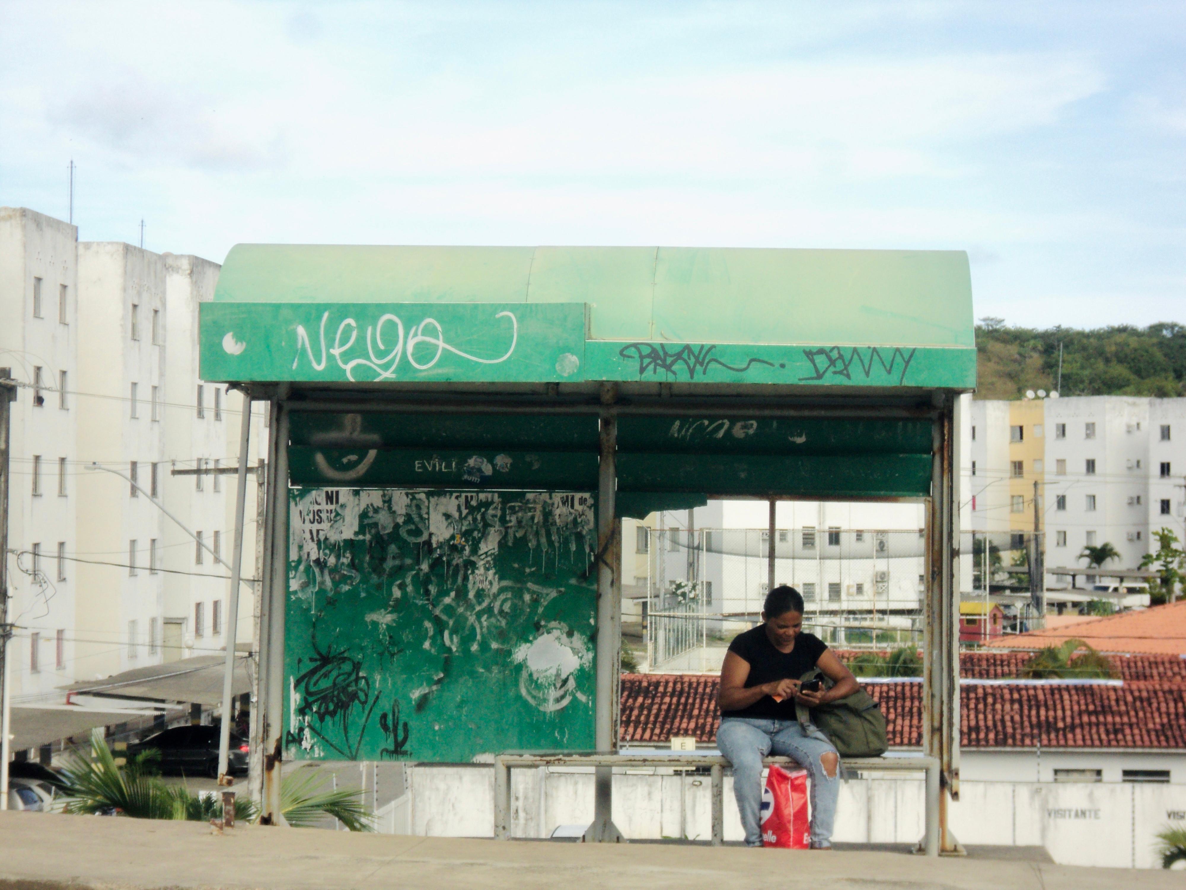 A imagem mostra um ponto de ônibus ao ar livre com uma pessoa sentada no banco. O ponto de ônibus tem um telhado e uma parede traseira verdes, ambos cobertos por várias pichações em diferentes cores. A pessoa está vestindo roupas escuras e olhando para algo em suas mãos, possivelmente um dispositivo móvel. Há uma bolsa vermelha ao lado da pessoa no banco. Ao fundo, há prédios brancos e árvores sob um céu nublado.