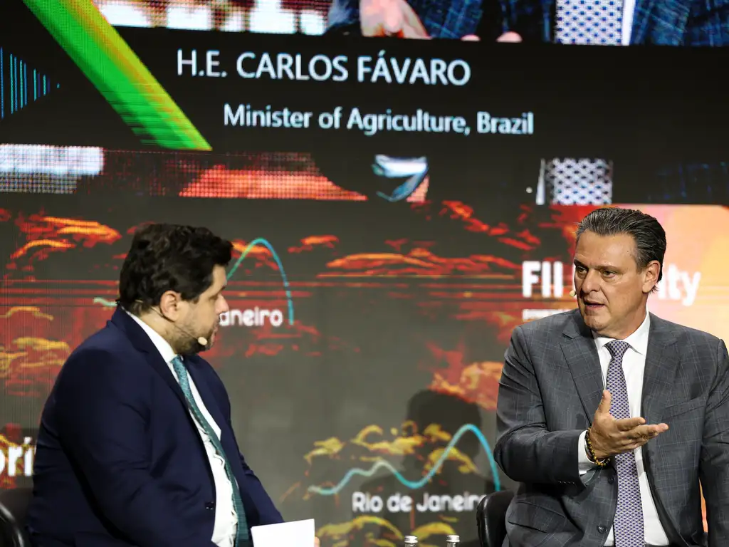 Foto de Carlos Fávaro, homem branco, de meia idade, com cabelos lisos escuros penteados para trás, usando paletó cinza, camisa branca e gravata cinza com grafismos brancos. Ele está em uma espécie de palco, tendo como interlocutor à esquerda da imagem, outro homem mais jovem, de cabelos escuros, mas que por estar de perfil não é possível ver completamente seu rosto. Os dois estão a frente de um grande painel onde é possível ler em inglês H.E. Carlos Fávaro Minister of Agriculture, Brazil.