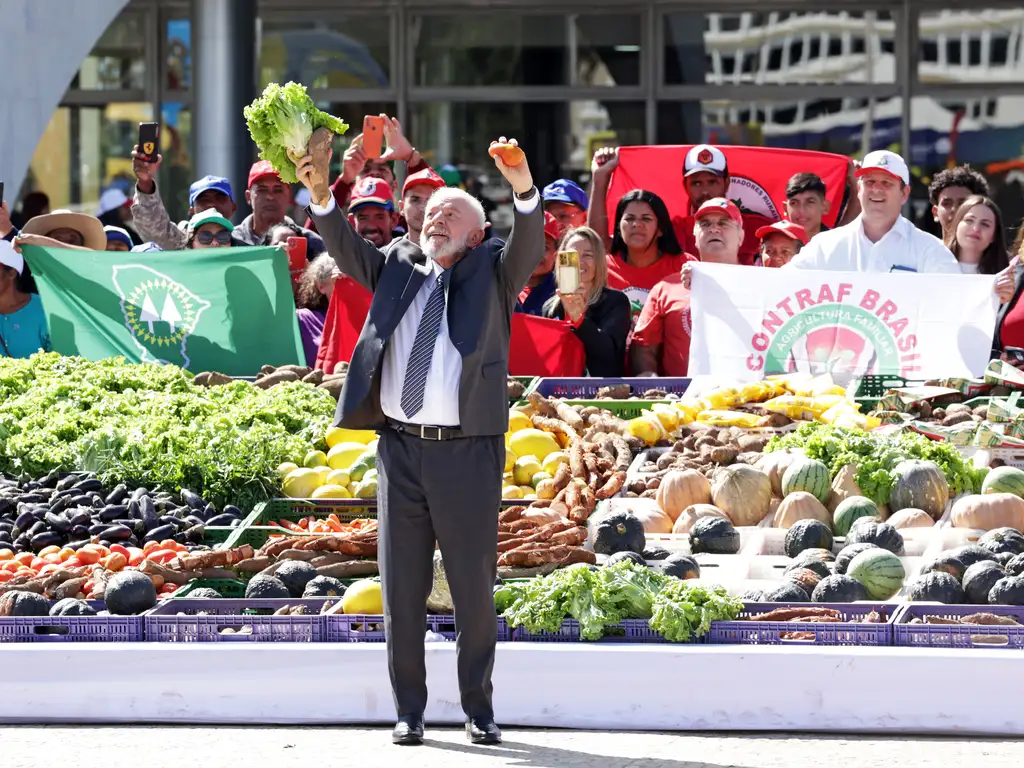 Foto do presidente Lula - homem idoso, de barba branca e cabelos ralos também brancos - está em pé, com os braços erguidos, segurando hortaliças na mão direita, à frente de uma vegetais dispostos em uma bancada junto ao chão. Por trás, da bancada de frutas e verduras, há um grupo de homens e mulheres segurando faixas e bandeiras de organizações ligadas à agricultura familiar e aos camponeses. A foto foi feita durante o dia e ao ar livre, no meio de uma rua ou pátio.