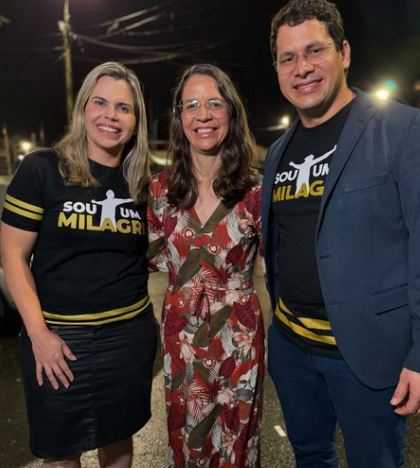 Foto noturna, ao ar livre, de Clarissa Tércio, Gil Tércio e Júnior Tércio. Clarissa é uma mulher loira, de cabelos lisos, usando saia preta e uma camiseta preta com detalhes dourados onde se lê a frase sou um milagre. Gil é uma mulher de cabelos castanhos compridos e ondulados abaixo dos ombros, usando um vestido estampado onde predomina as cores vermelho e marrom; Júnior é um homem alto, branco, de cabelos vastanhos curtos, óculos, usando uma camiseta igual a de Clarissa, porém com um terno azul e calça da mesma cor.