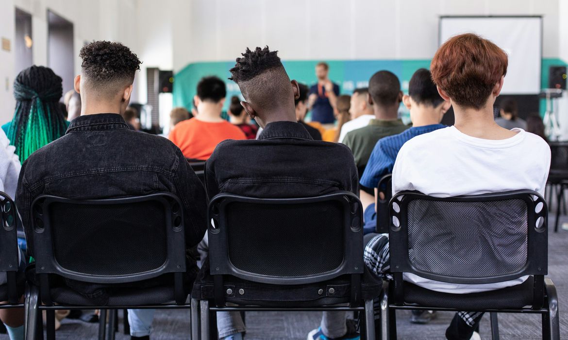 A imagem mostra um grupo de jovens, negros e brancos, sentados em cadeiras de plástico preto, vistas de costas. As cadeiras estão dispostas em fileiras, e as pessoas parecem estar assistindo a uma apresentação ou palestra. Há uma pessoa em pé na frente, um pouco desfocada, possivelmente falando ou apresentando algo. Ao fundo, há um projetor com uma tela branca. A sala parece ser um ambiente educacional ou de conferência, com várias pessoas prestando atenção ao que está sendo apresentado.