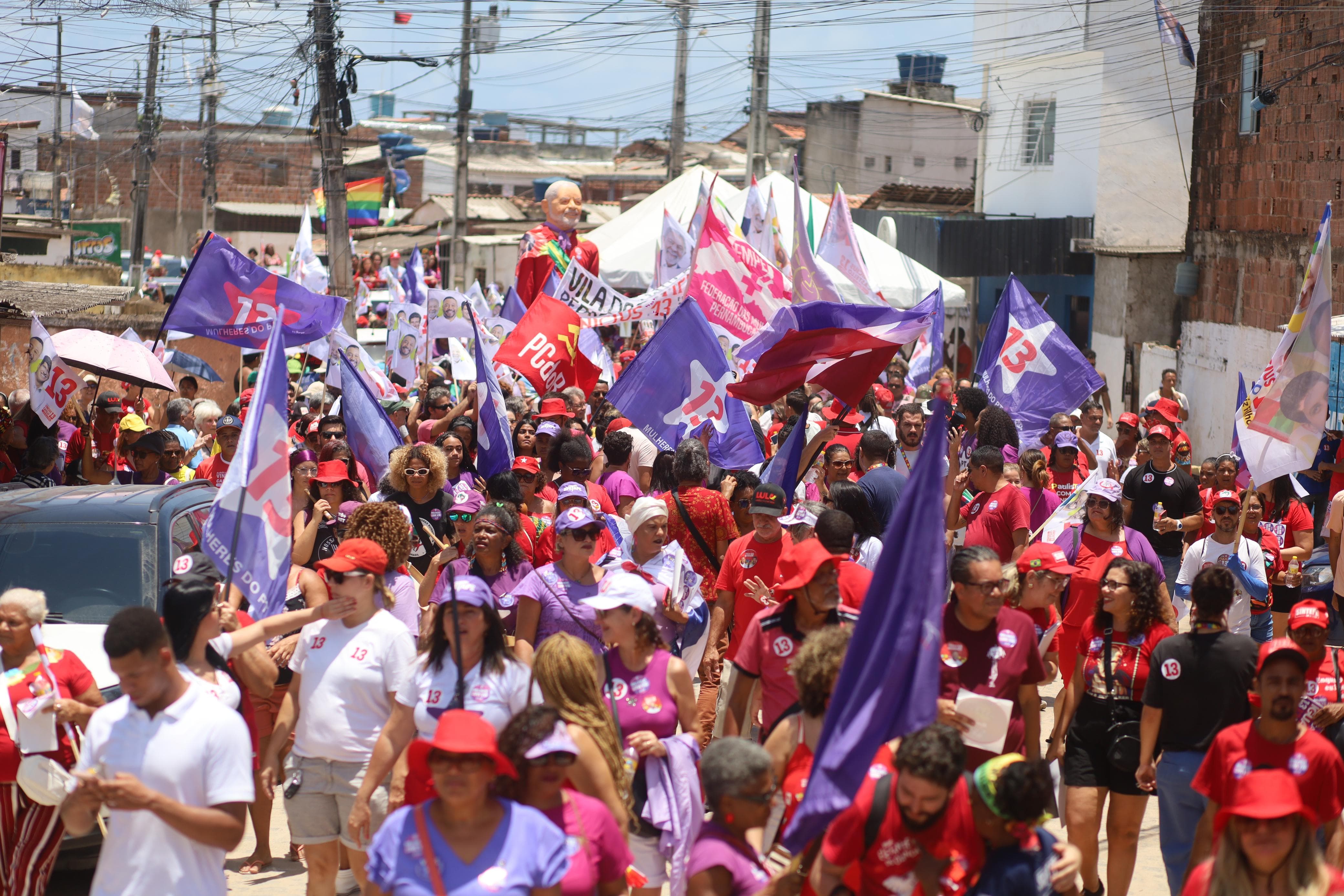 Foto colorida de uma multidão no meio da rua, com a maioria das pessoas vestindo camisetas vermelhas ou brancas, além de portarem bandeiras na cor lilás com estrela branca e o número 13 na cor vermelha.