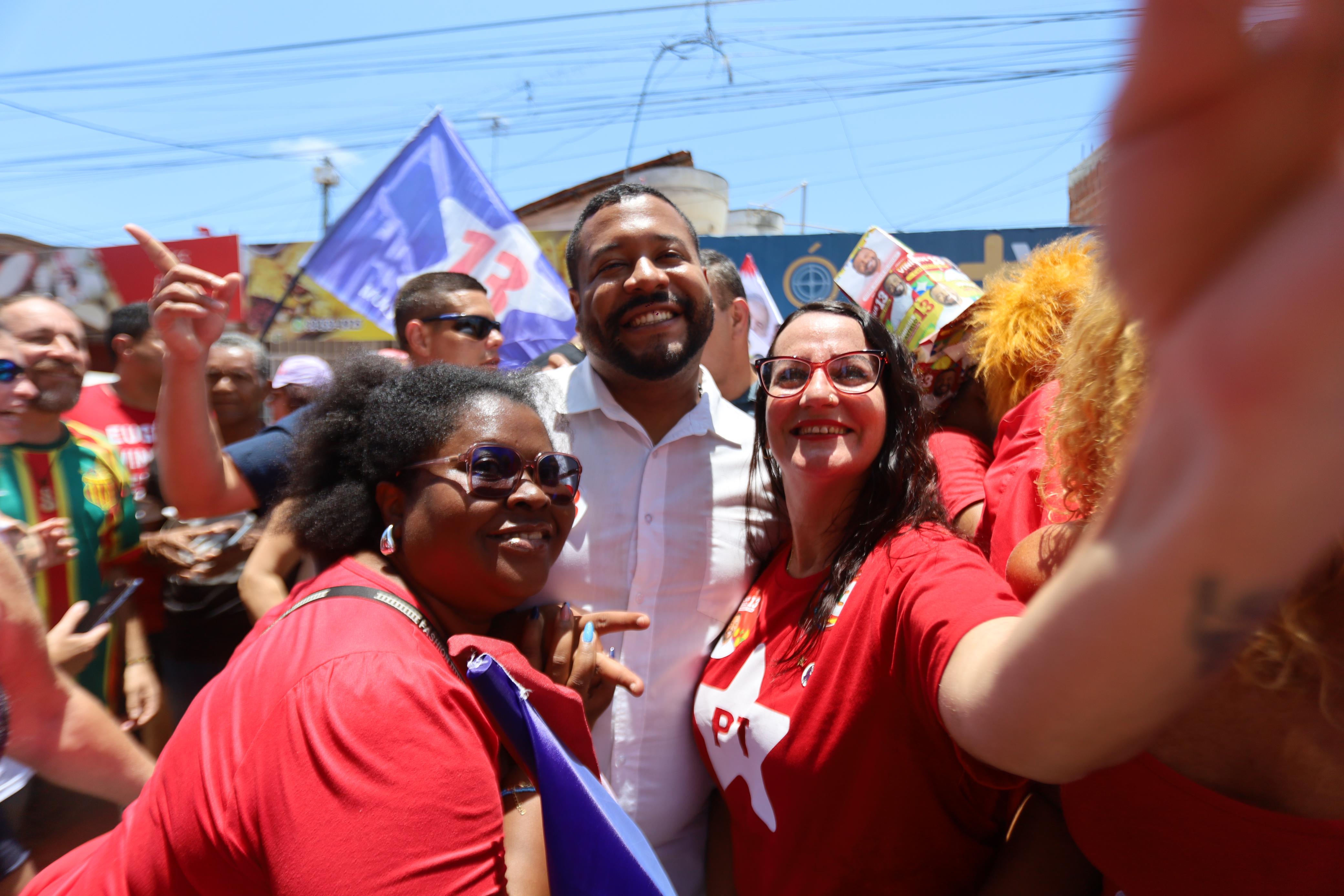 Foto colorida de Vinicius Castelo no meio da rua, cercado por dezenas de pessoas, olhando e sorrindo para a câmera entre duas mulheres. Ele é um homem negro, jovem, de cabelos curtos, vestindo uma camisa branca de botões. A mulher à esquerda da imagem é uma mulher negra, jovem, de cabelos crespos, usando camiseta vermelha e óculos escuros; a mulher à direita tem pele clara, cabelos pretos escorridos, com óculos de grau de armação vermelha e veste camiseta vermelha estampada com estrela branca do PT e, com sua mão esquerda, aparenta estar segurando um celular para fazer uma selfie.