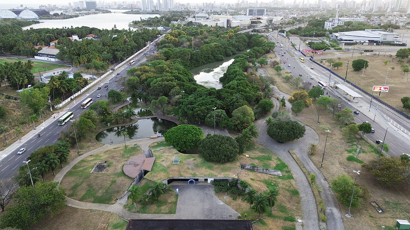 A imagem mostra vista aérea do Espaço Ciência, um parque urbano com áreas de vegetação, lagos e caminhos pavimentados. À esquerda, há uma avenida movimentada e, ao fundo, vê-se prédios altos e um corpo d'água. O parque tem espaços gramados, árvores e pequenas estruturas. À direita, outra avenida margeia o local. A cena mistura natureza e urbanização.